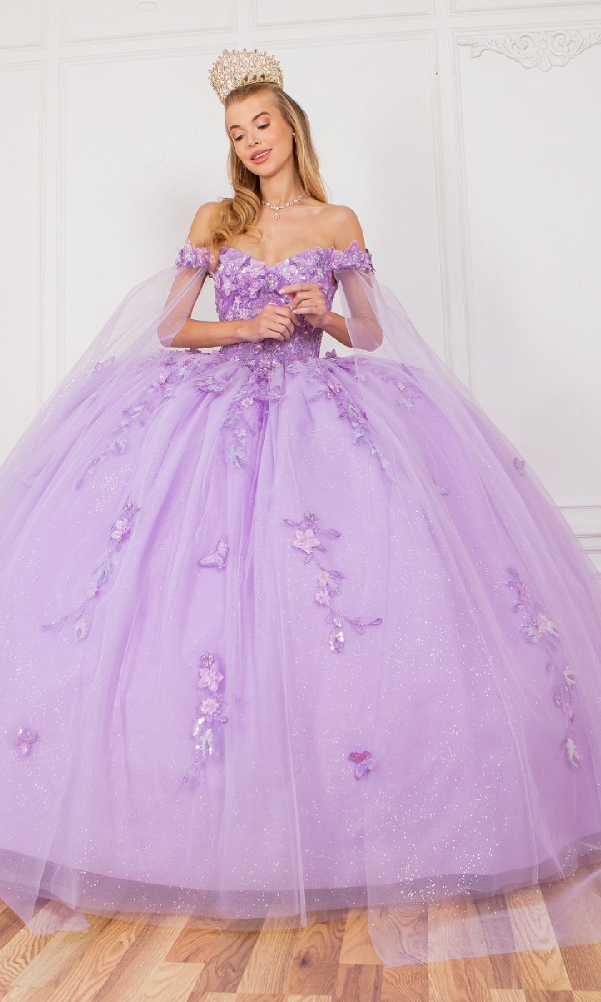 A woman stands indoors on a wooden floor wearing the Cinderella Couture 8075J quinceanera dress in glittery lavender with 3D floral appliques, a detachable sheer cape, and a gold crown.